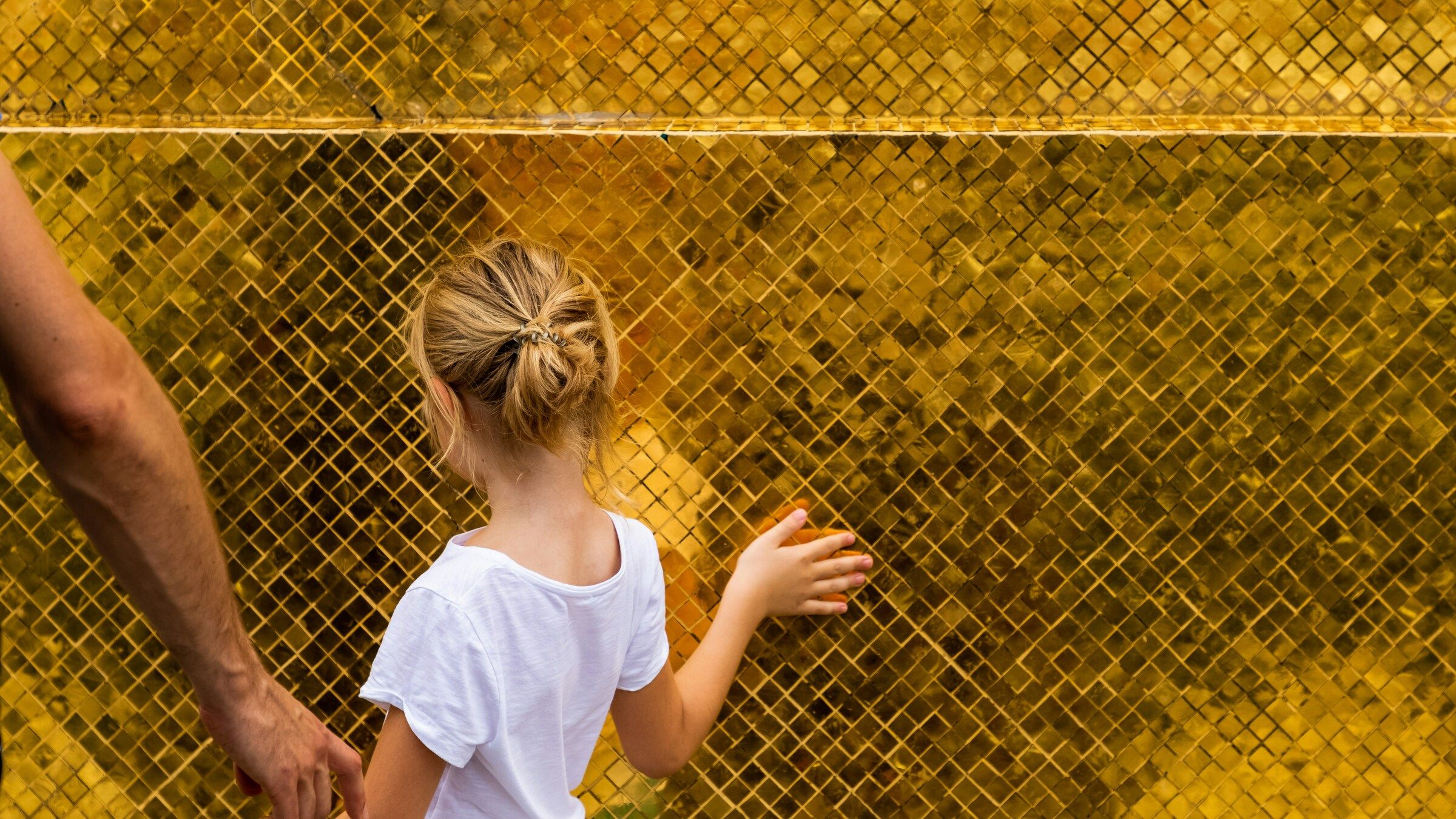 Seen from behind, a child holding a parent's hand touches a golden mosaic wall in warm light, with no face shown.