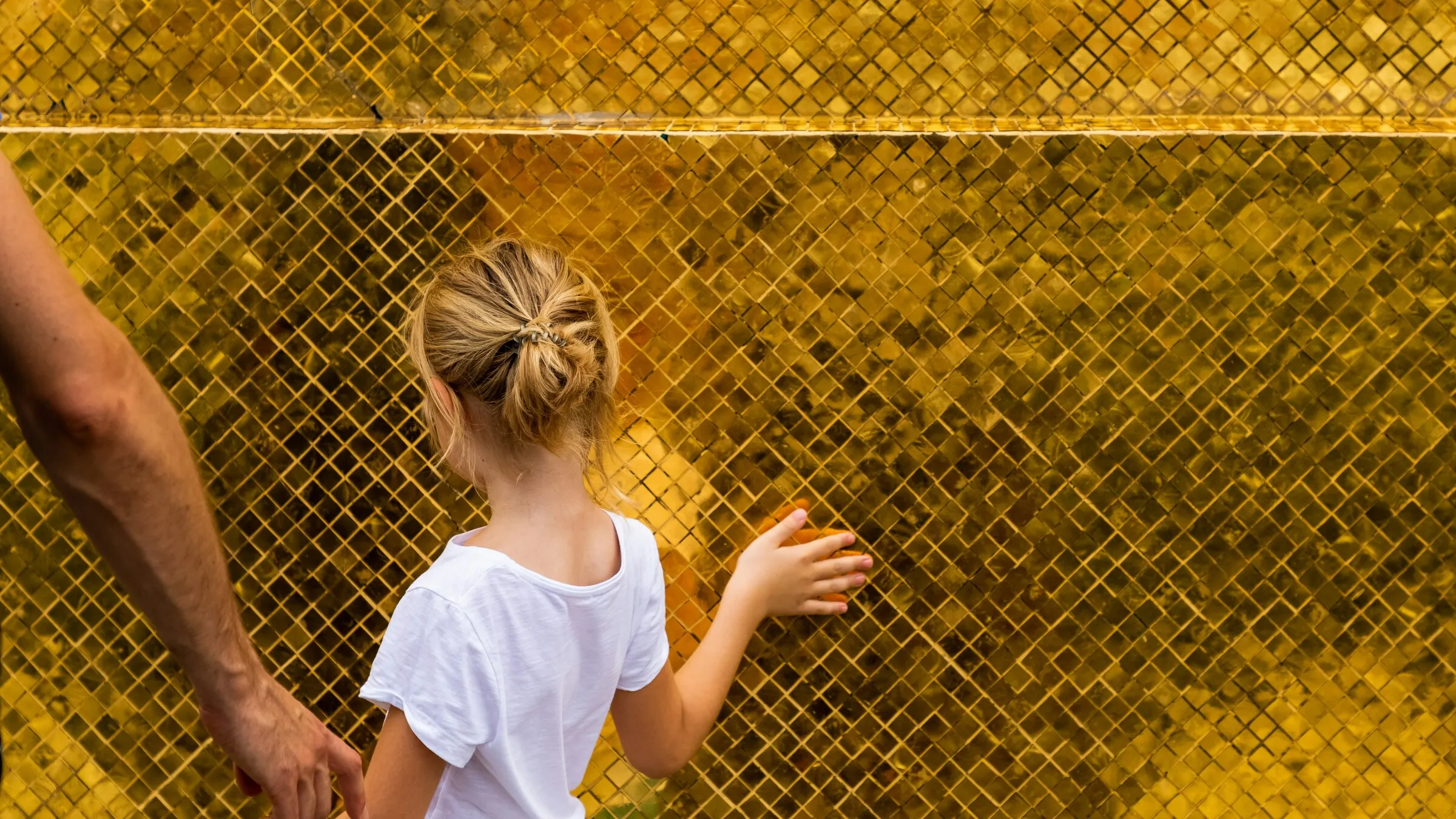 Seen from behind, a child holding a parent's hand touches a golden mosaic wall in warm light, with no face shown.