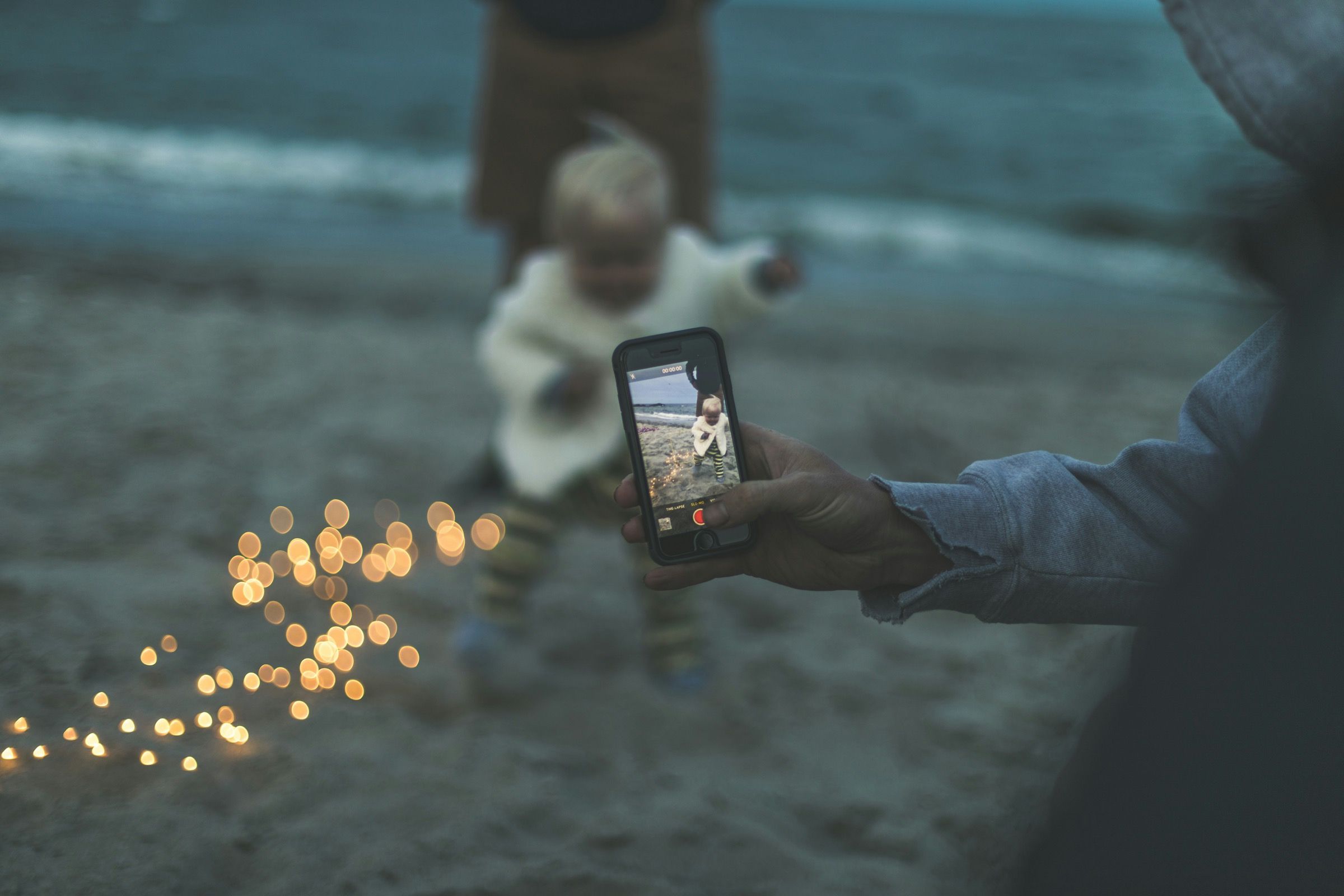 Close-up of a parent's hand filming their toddler on a phone at a beach at dusk, the child's face out of focus, with warm bokeh lights.